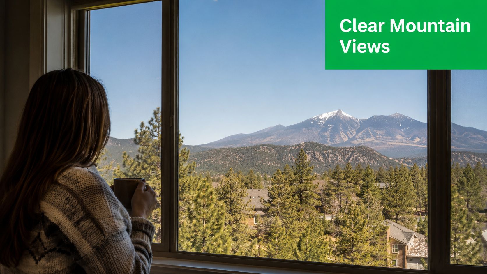 A woman stands by a window holding a coffee mug looking out at snowy mountains.